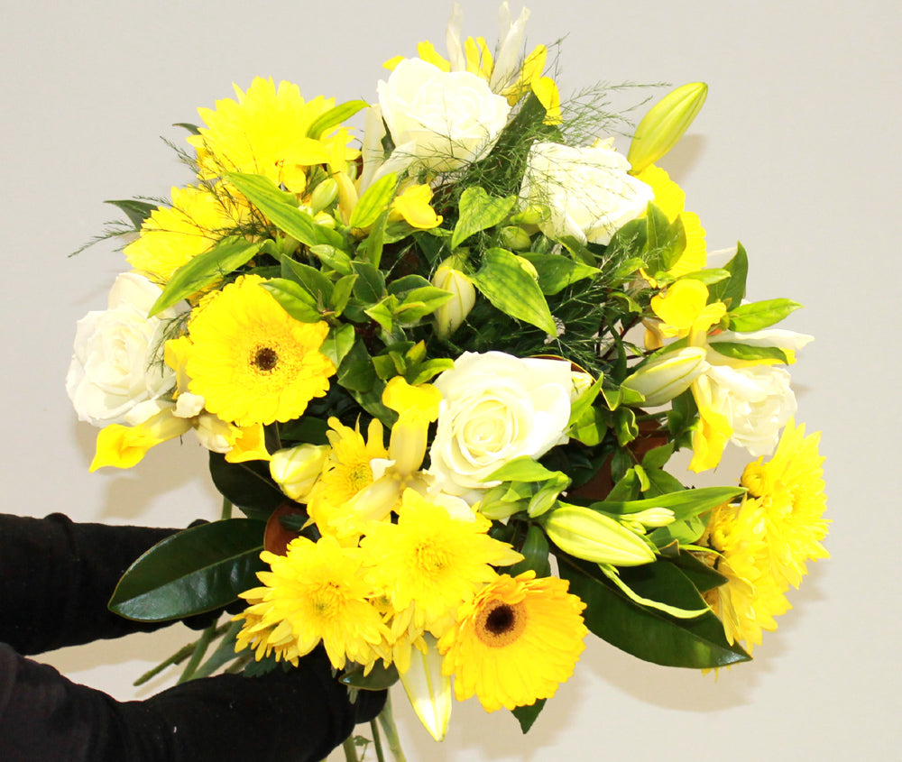 A vibrant flower bouquet featuring yellow and white flowers and green foliage, held by the florist