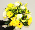A vibrant flower bouquet featuring yellow and white flowers and green foliage, held by the florist
